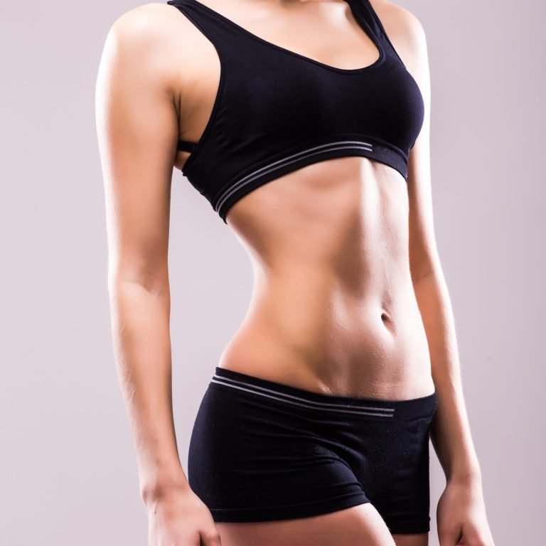 Muscular young woman athlete standing looking down with her hands on hips on grey background. Woman bodybuilder relaxing after exercise.